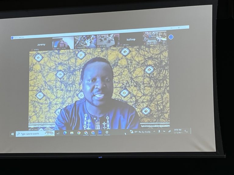 Two computer screens displaying a video call with a man in a patterned shirt against a yellow background.