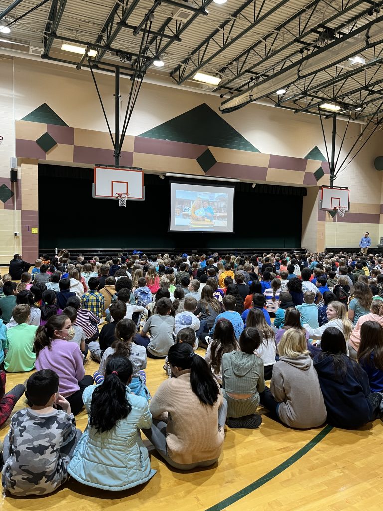 A group of children seated on the floor in a gymnasium watching a presentation on a projector screen.