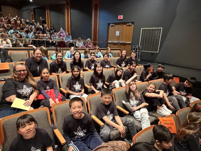 People sitting in a theater, all wearing black shirts with white designs. They are posing for a picture.