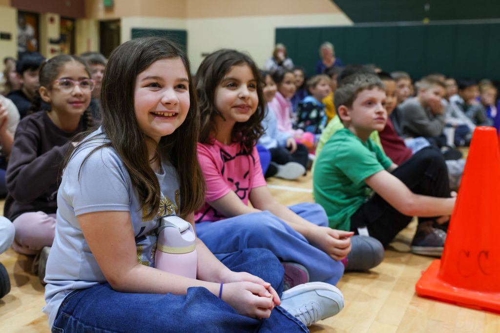 Children sitting on gym floor, watching event through window.