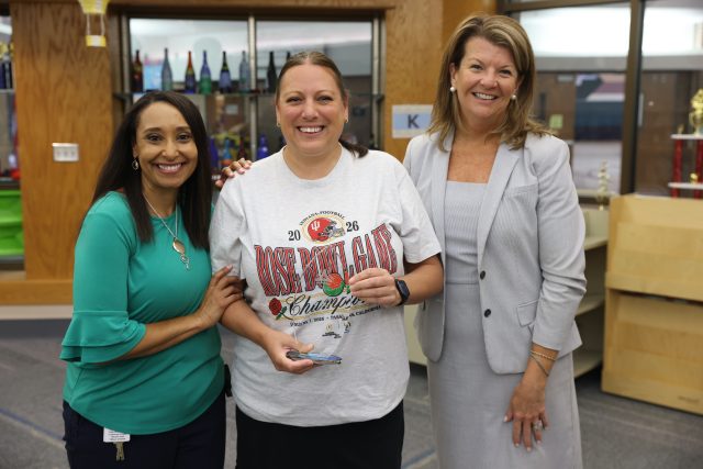 Three individuals posing together in an office setting, one woman holding a trophy.